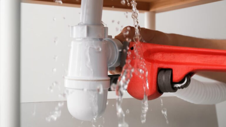 A red wrench clasps onto a pipe under a sink, water falls all around it.