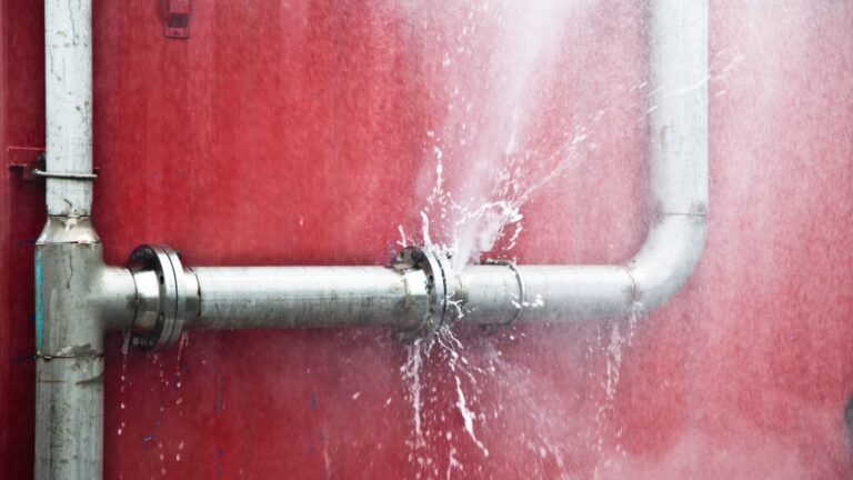 A silver pipe along a red wall bursts with water, the water sprays in all directions.